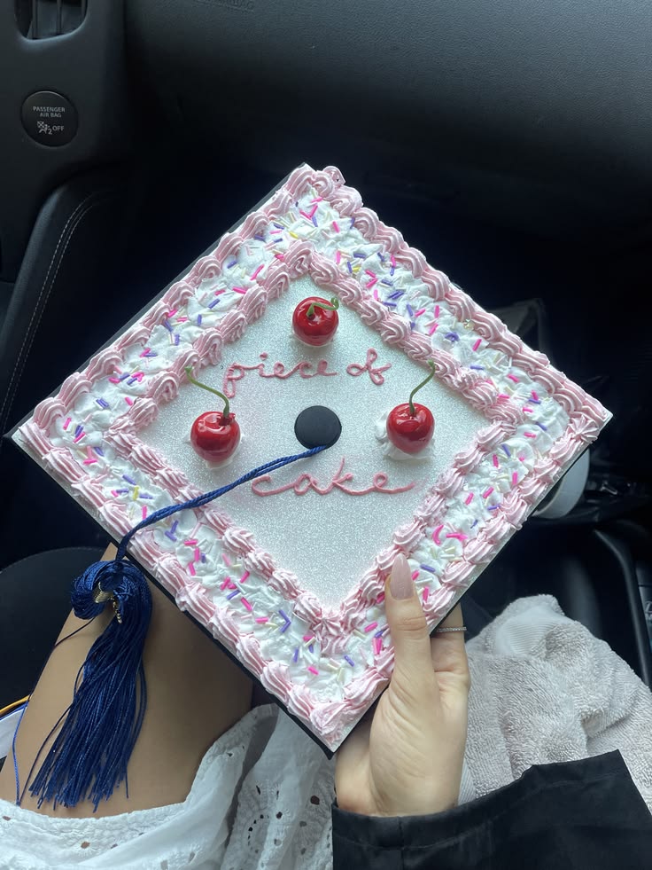 Graduation cap decorated to look like a birthday cake with pink frosting swirls, rainbow sprinkles, and red cherries with the words piece of cake written in the centre.