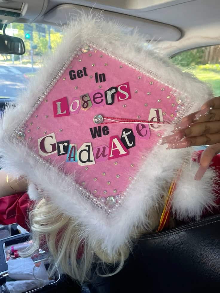 Pink graduation cap trimmed with white fluffy marabou feathers, rhinestones, and cut-out ransom-style letters spelling get in losers we graduated.