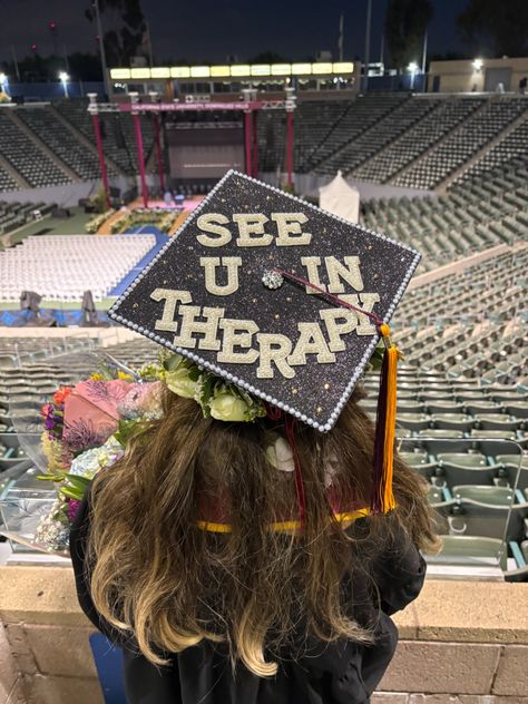 Dark navy glitter graduation cap with gold glitter letters spelling see u in therapy, decorated with a rhinestone brooch and pearl border.
