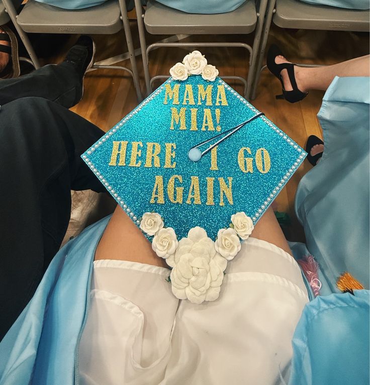 Teal blue glitter graduation cap with gold glitter letters reading Mama Mia here I go again, decorated with white floral roses.