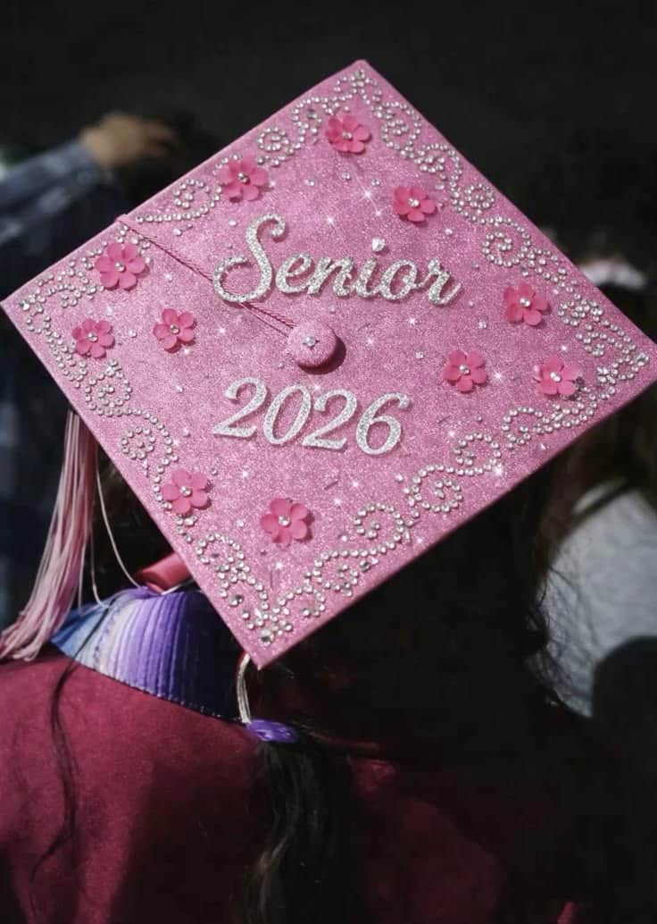 Pink glitter graduation cap with silver rhinestone swirl designs, small pink paper flowers, and silver glitter lettering reading Senior 2026.