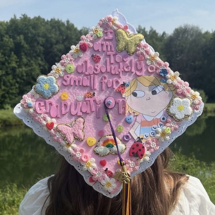 Pink graduation cap heavily decorated with piped rosette trim, paper flowers, butterflies, ladybugs, buttons, and a hand-drawn cartoon character with the words I am too absolutely small for graduation.