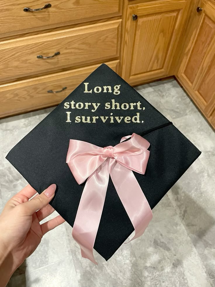 Black graduation cap with gold serif text reading long story short I survived, finished with a large pink satin bow.