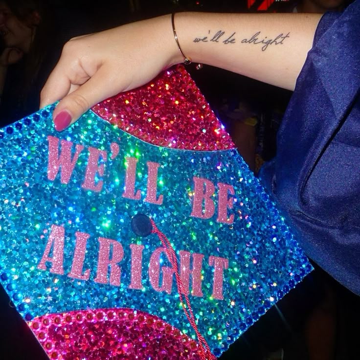 Blue and pink holographic glitter graduation cap with pink glitter letters spelling we'll be alright.