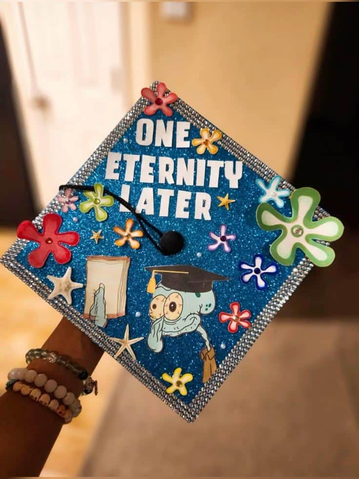 Blue glitter graduation cap with rhinestone border featuring a printed Squidward wearing a graduation cap, SpongeBob flowers, and the words one eternity later in white.