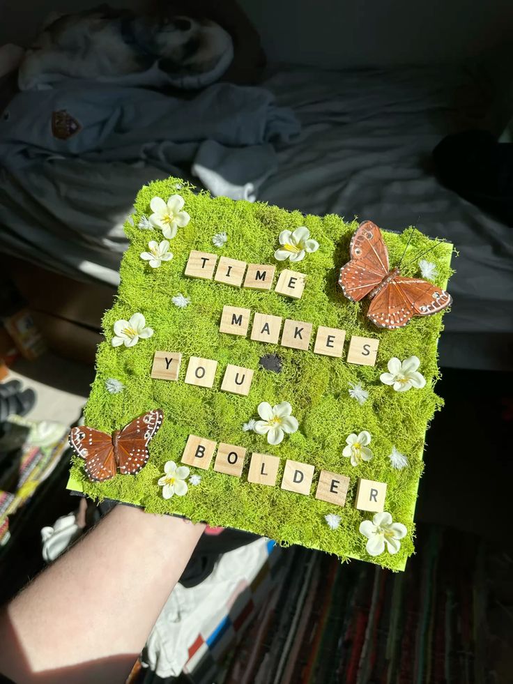 Graduation cap covered in green moss with Scrabble tile letters spelling time makes you bolder, decorated with white flowers and two rust coloured butterfly embellishments.