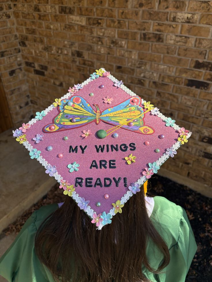 Pink glitter graduation cap with a large colourful butterfly in the centre, pastel paper flowers along the edges, and black text reading my wings are ready.