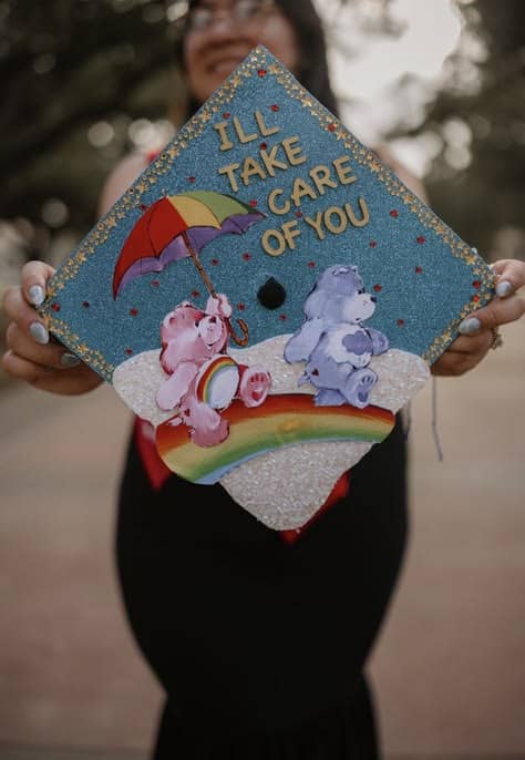 Teal blue glitter graduation cap with Care Bears characters on a rainbow, gold glitter letters reading I'll take care of you, and a rhinestone star border.