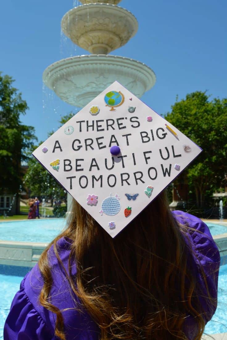 White graduation cap with black handwritten text reading there's a great big beautiful tomorrow, decorated with colourful small sticker charms including a globe, flowers, butterfly, disco ball, and strawberry.