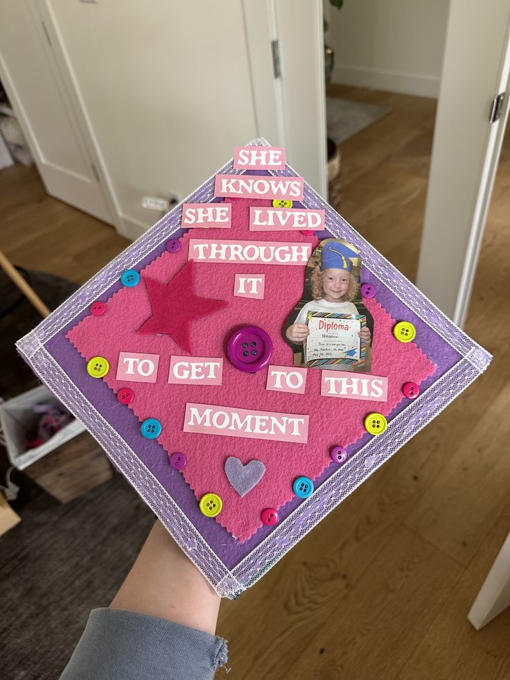 Purple and pink graduation cap with white letter stickers, colourful buttons, a felt star, and a childhood photo of the graduate holding a preschool diploma.