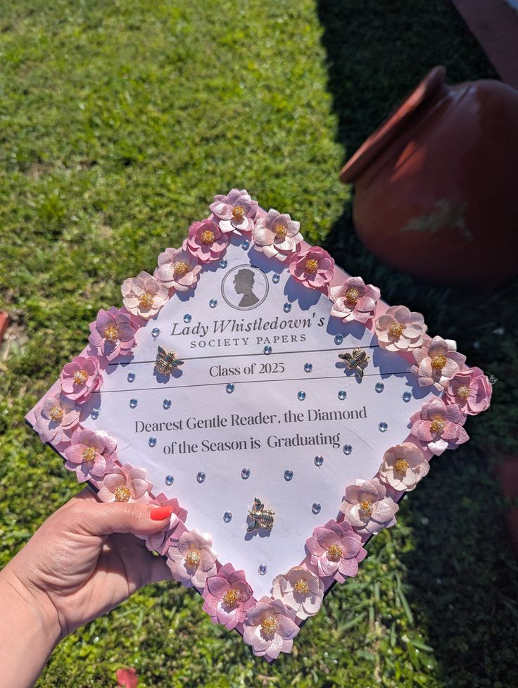 White graduation cap styled as a Lady Whistledown Society Papers announcement bordered with pink paper flowers and rhinestones, reading Dearest Gentle Reader the Diamond of the Season is Graduating