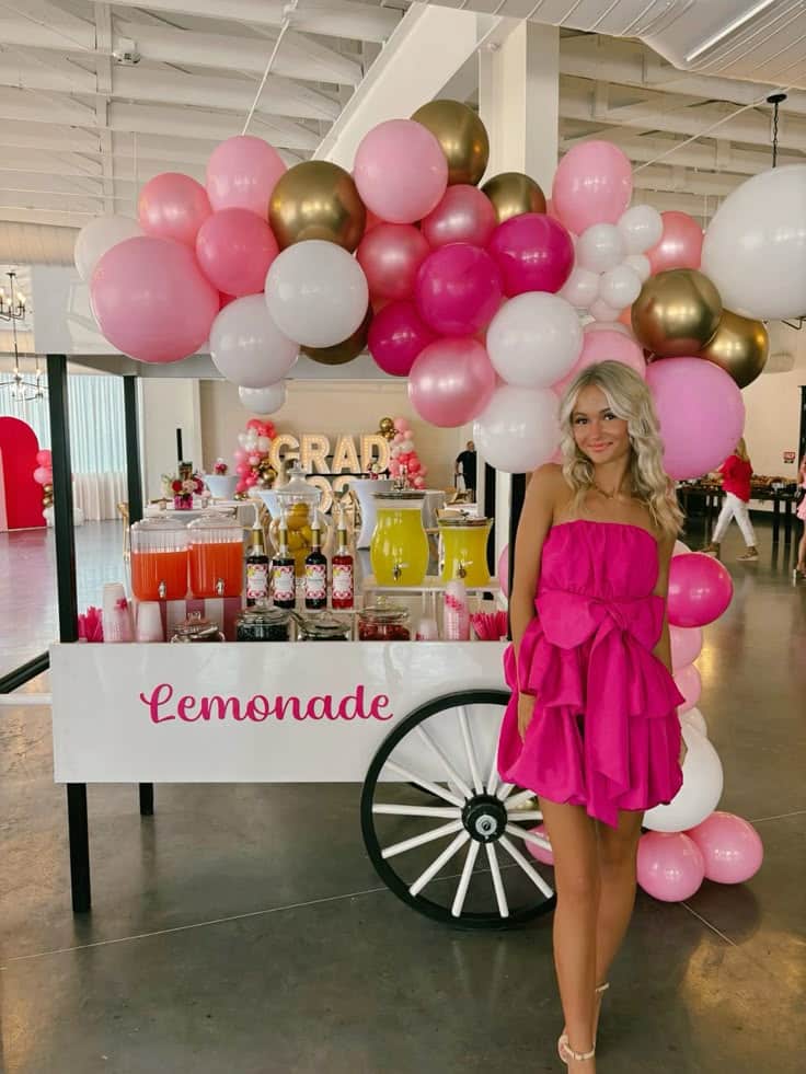 Girl in pink dress standing next to a white lemonade cart with colourful drink dispensers and a pink and gold balloon garland above at a graduation party