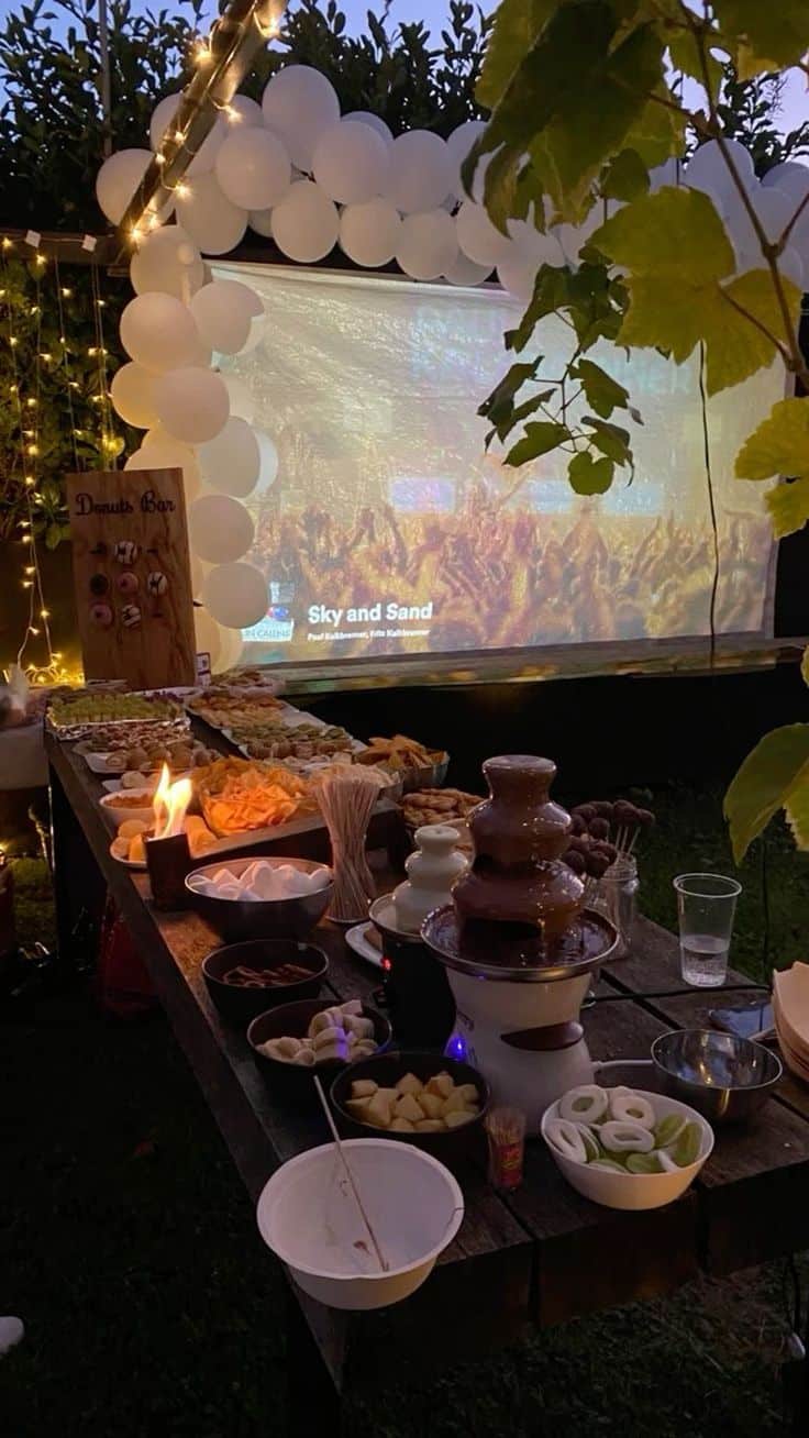 Evening outdoor graduation party setup with a projector screen a chocolate fountain surrounded by dipping snacks and a white balloon arch with fairy lights