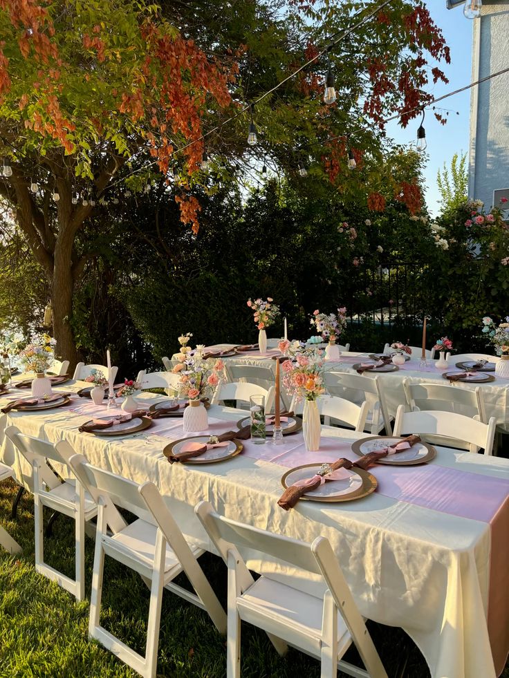 Outdoor graduation party dining tables with cream and pink linens gold charger plates fresh floral centrepieces and Edison bulb string lights overhead