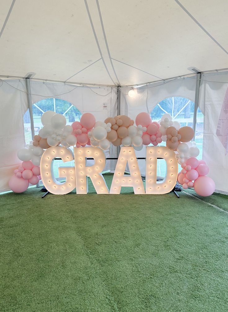 Large illuminated white marquee letters spelling GRAD with a pink white and peach balloon garland inside a marquee tent at a graduation party