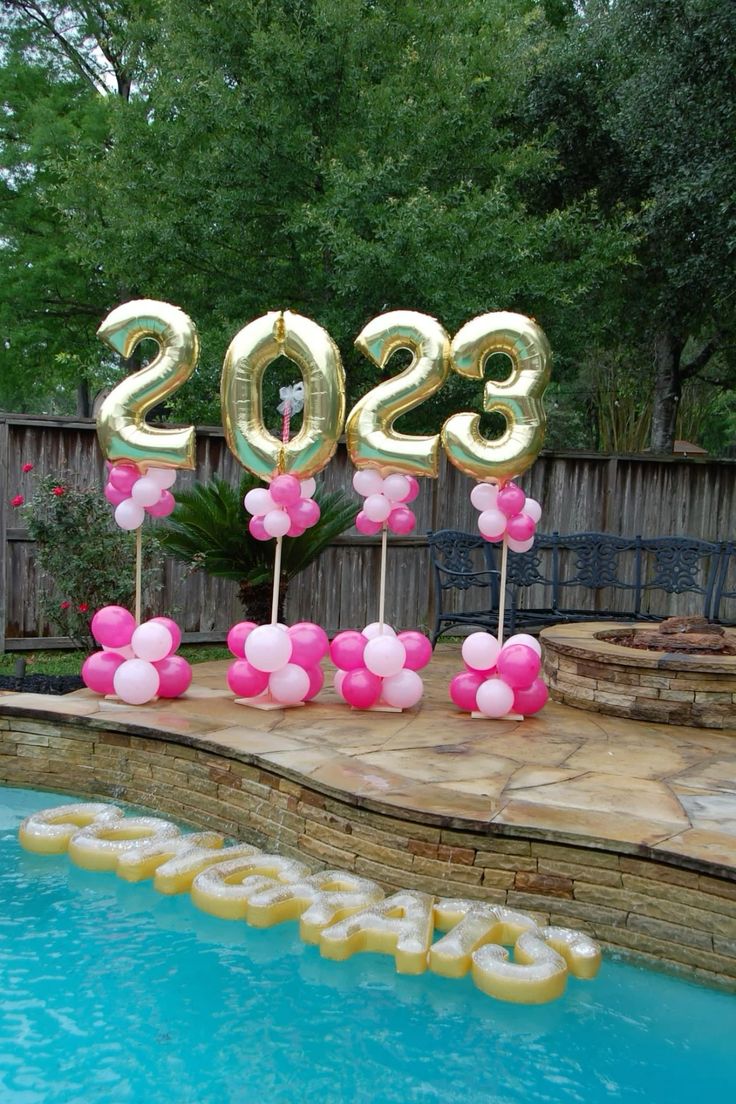 Gold foil graduation year number balloons on pink balloon stands beside a pool with floating Congrats letters in the water