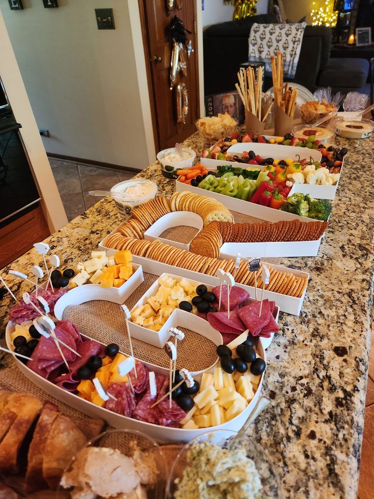 Graduation party grazing table with charcuterie meats cheeses crackers olives and a vegetable platter spread across a kitchen counter