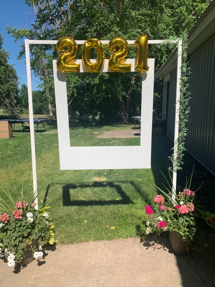 DIY white giant polaroid photo frame standing outdoors with gold graduation year balloons on top and flower pots at the base