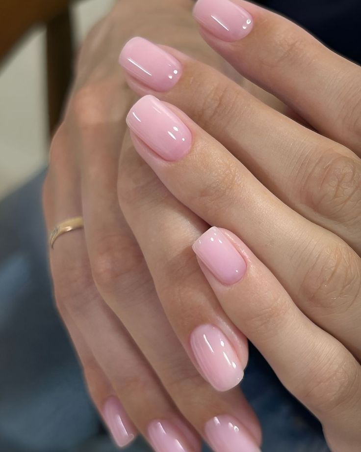 Close-up of short square nails painted in a soft, glossy milky pink gel on interlaced hands wearing a gold ring