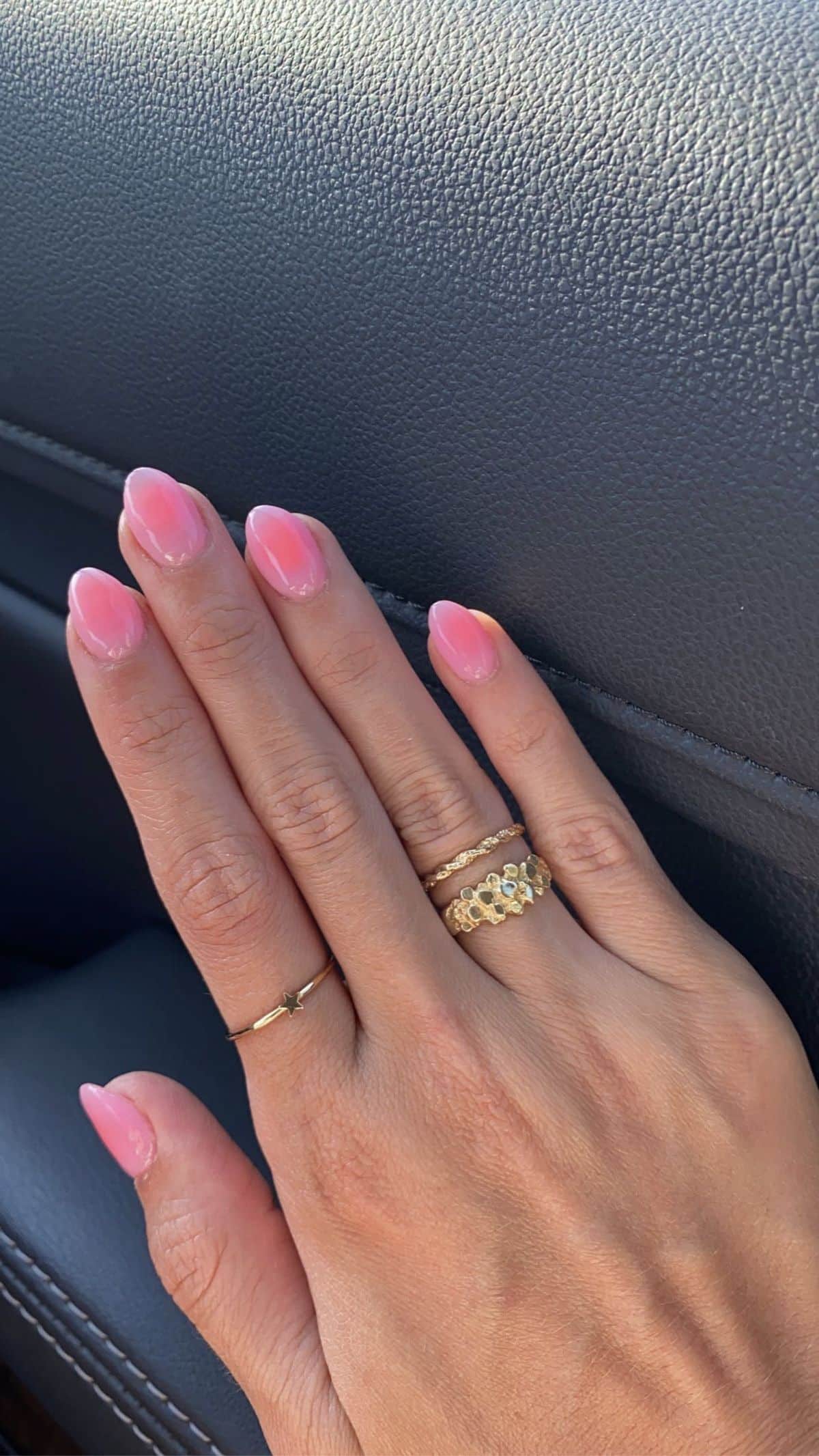 Medium oval nails with a soft blush pink ombre fading into a sheer natural base, on a hand wearing gold stacked rings, resting on a car seat