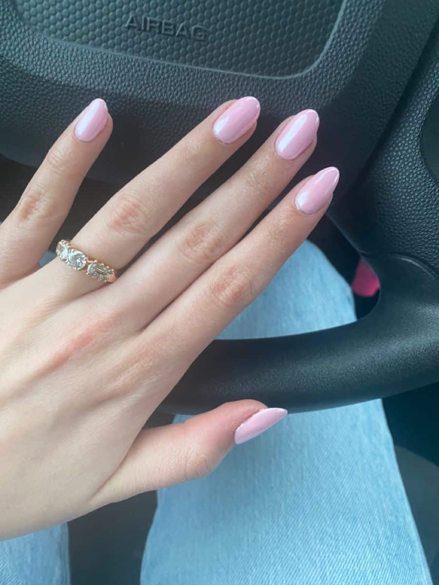 Medium almond nails with a light glossy pink gel finish on a hand wearing a multi-stone gold ring, resting on a car steering wheel in denim