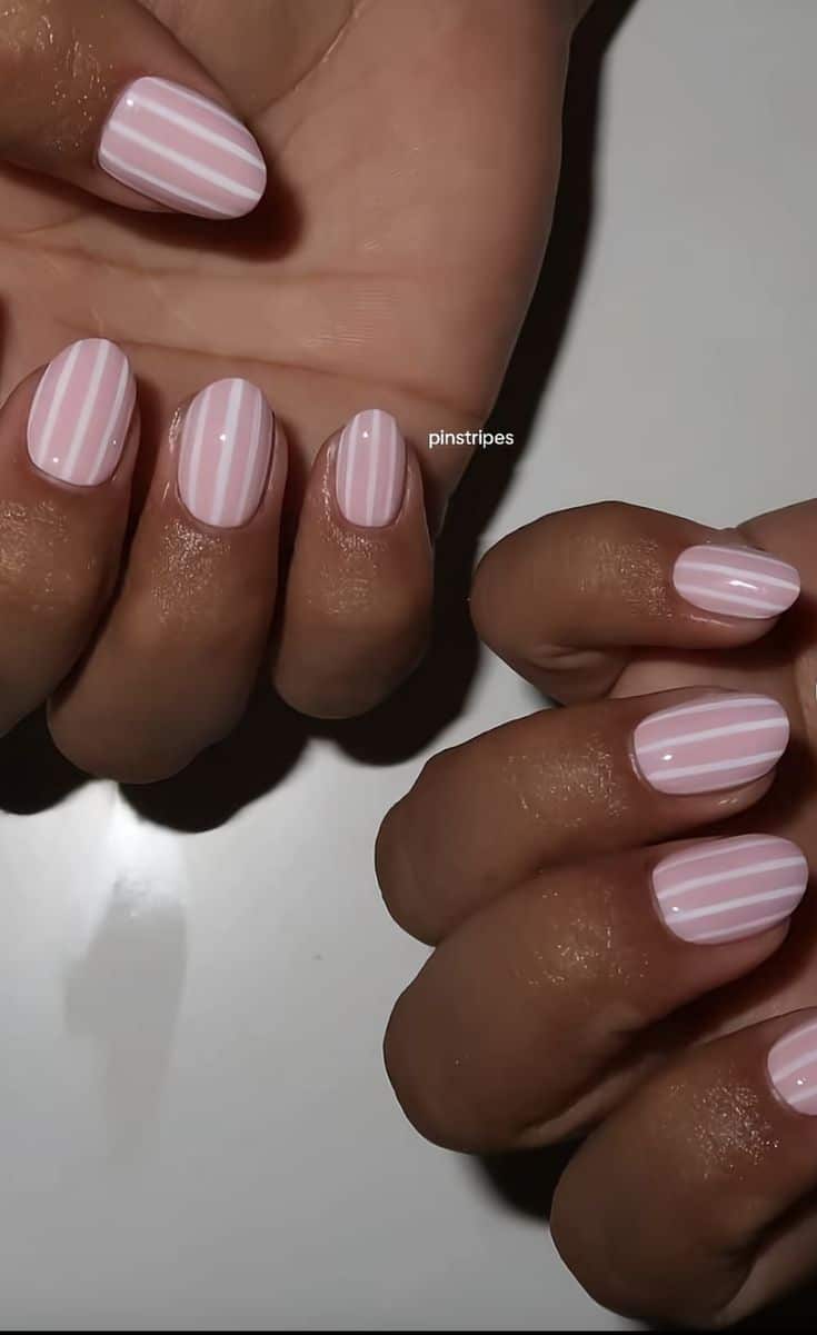 Short rounded square nails in a light pink gel with white vertical pinstripes on two sets of hands resting on a white surface