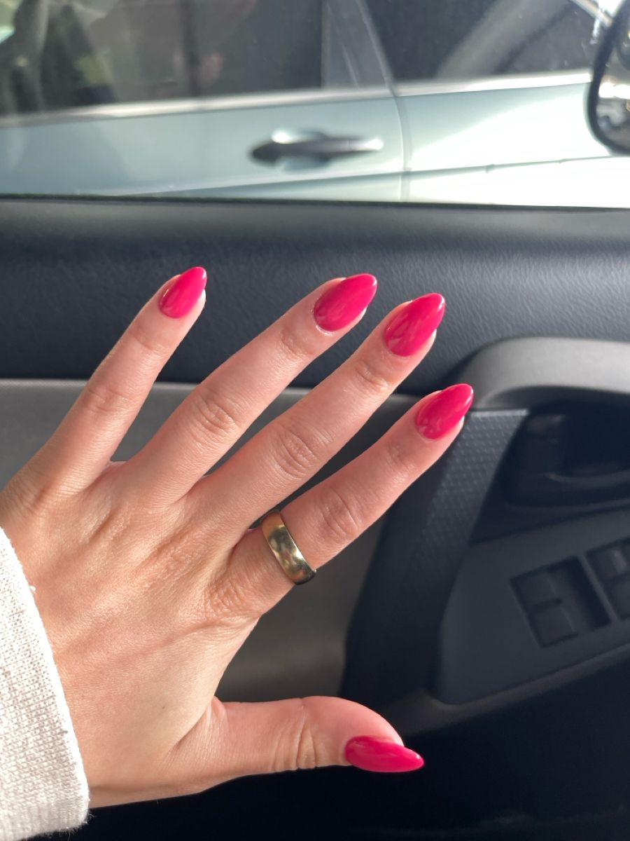 Long almond nails in a deep warm raspberry pink gel on a hand with a gold wedding band, resting against a car door panel