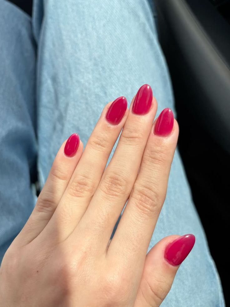Medium almond nails in a deep berry pink gel on a hand resting on a denim lap with a car in the background