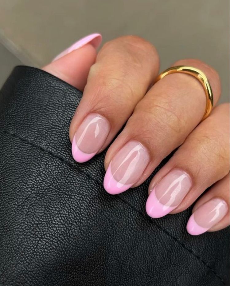 Medium almond nails with a sheer natural base and a soft bubblegum pink curved French tip, on a hand wearing a gold ring against a dark leather background