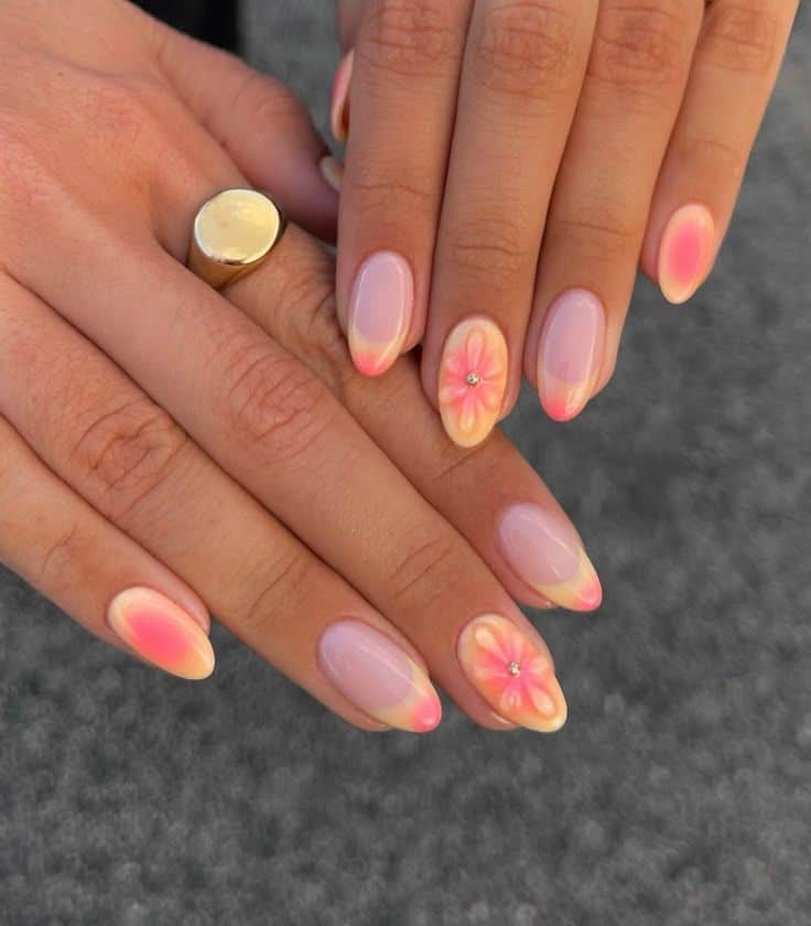 Medium almond nails with a sheer base, soft pink and peach aura blush spots, and hand-painted coral daisy flowers with rhinestone centres on two hands wearing a gold signet ring