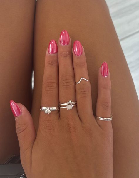 Almond-shaped nails with a glossy hot pink chrome mirror finish on a tanned hand wearing silver rings, resting on a leg