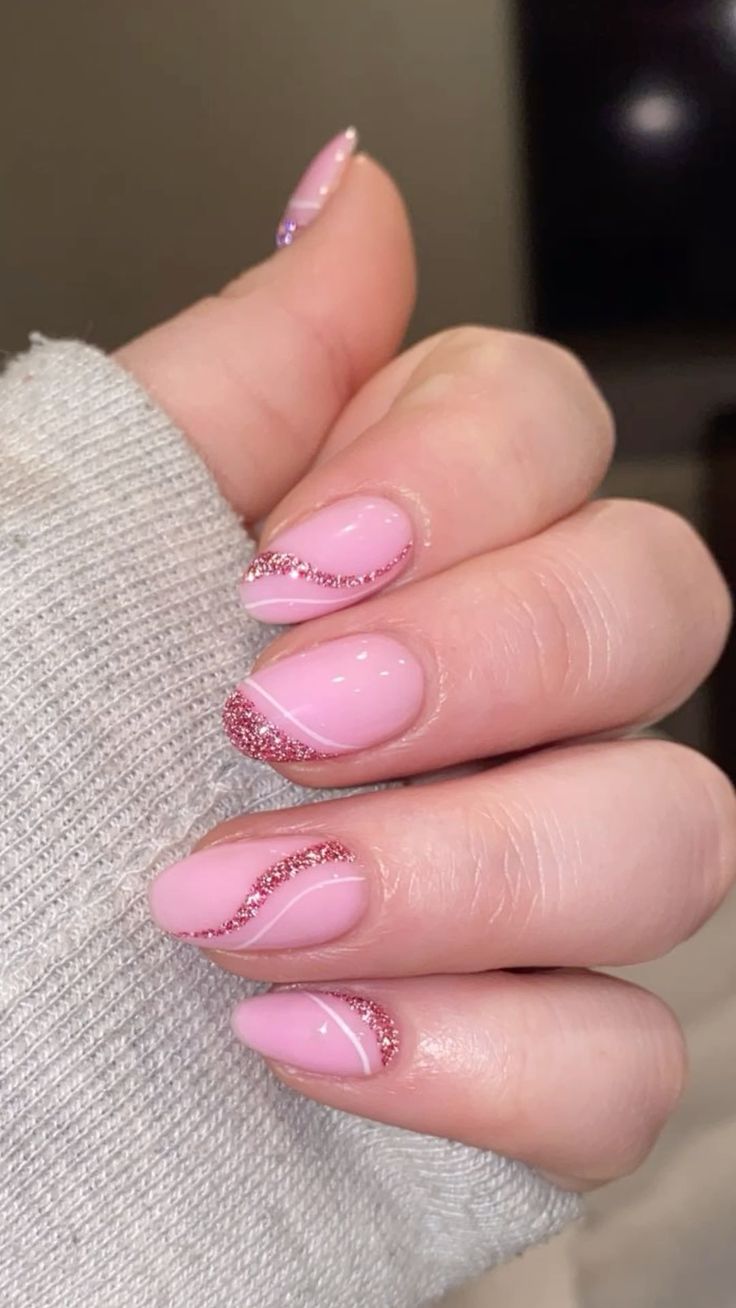 Medium oval nails in a soft pink gel with a diagonal sweep of pink chunky glitter across each nail, on a hand in a cream knit sleeve