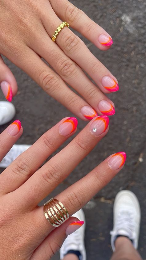 Short oval nails with a sheer natural base and bright neon pink and orange abstract double French tips, on two hands wearing gold rings, photographed outdoors