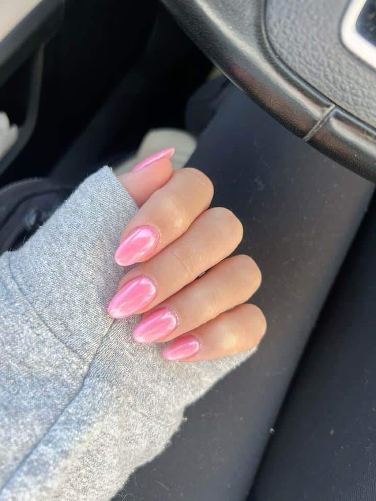 Medium almond nails with a warm pink chrome pearlescent finish on a hand resting on a grey car seat wearing a grey sweatshirt