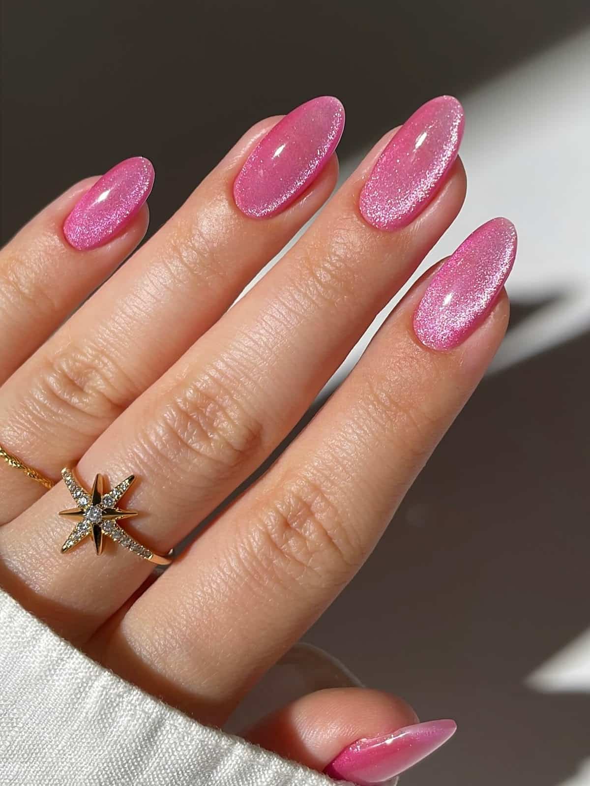 Medium almond-shaped nails with a warm rosy pink gel and fine shimmer finish, on a hand wearing a gold starburst ring against a light background