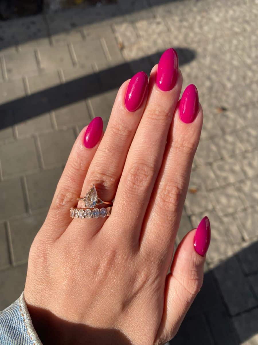 Long almond nails painted in a vivid hot pink gel on a hand wearing a pear-shaped diamond ring and diamond eternity band, photographed outdoors on cobblestones