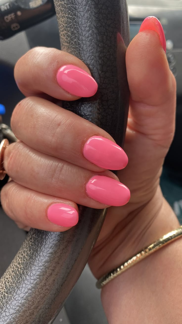 Medium almond nails in a bright warm bubblegum pink gel on a hand gripping a car steering wheel, wearing a gold bangle