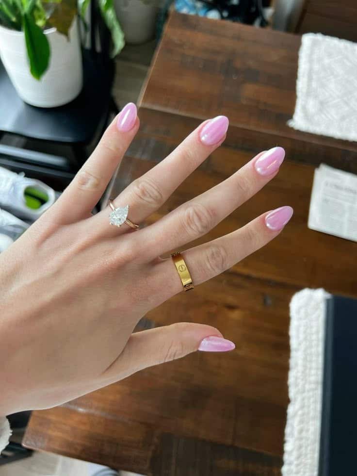 Medium oval nails with a soft pink chrome pearlescent finish on a hand wearing a pear-shaped engagement ring and a Cartier love ring