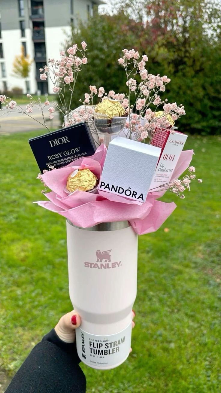 White Stanley Iceflow Flip Straw tumbler held outdoors filled with Ferrero Rocher chocolates, a Pandora box, Dior Rosy Glow blush, Clarins lip oil, and pink gypsophila