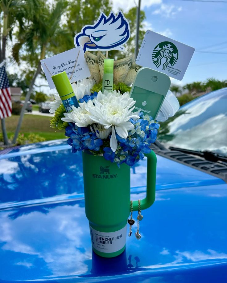 Green Stanley Quencher tumbler on a car hood filled with blue hydrangeas, white chrysanthemums, a Starbucks gift card, NYX lip product, Touchland sanitizer, cash, and a school mascot topper
