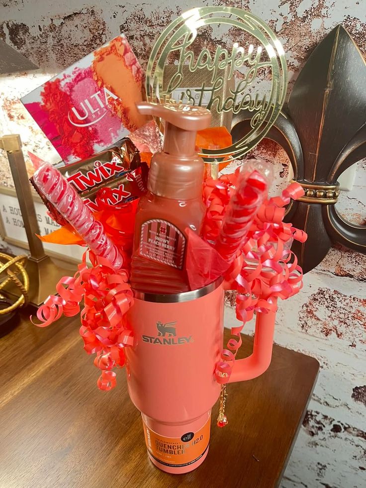 Coral orange Stanley Quencher tumbler decorated with matching curly ribbon, Bath and Body Works lotion, Twix chocolate bars, an Ulta palette, and a Happy Birthday acrylic topper