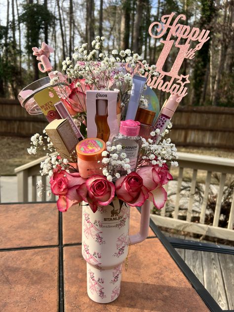 Floral-patterned Stanley tumbler outdoors filled with spray roses, baby's breath, Gisou hair oil, Garnier micellar water, Burt's Bees products, and a pink Happy 11th Birthday acrylic topper