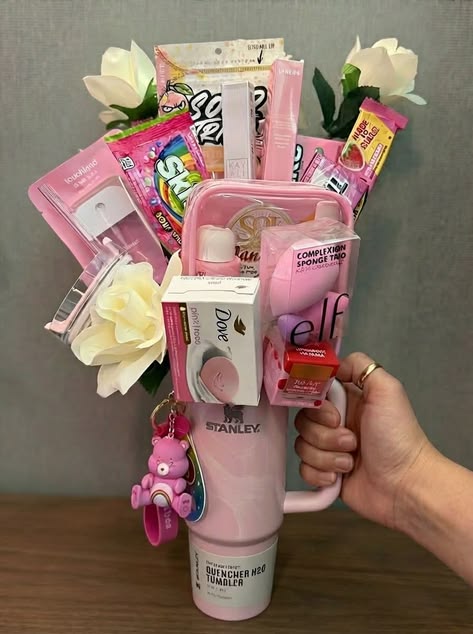 Pink Stanley Quencher tumbler held by hand with Care Bear keychain and filled with Kayali perfume, e.l.f. beauty products, Dove soap, Sol de Janeiro lotion, candy, and artificial white roses
