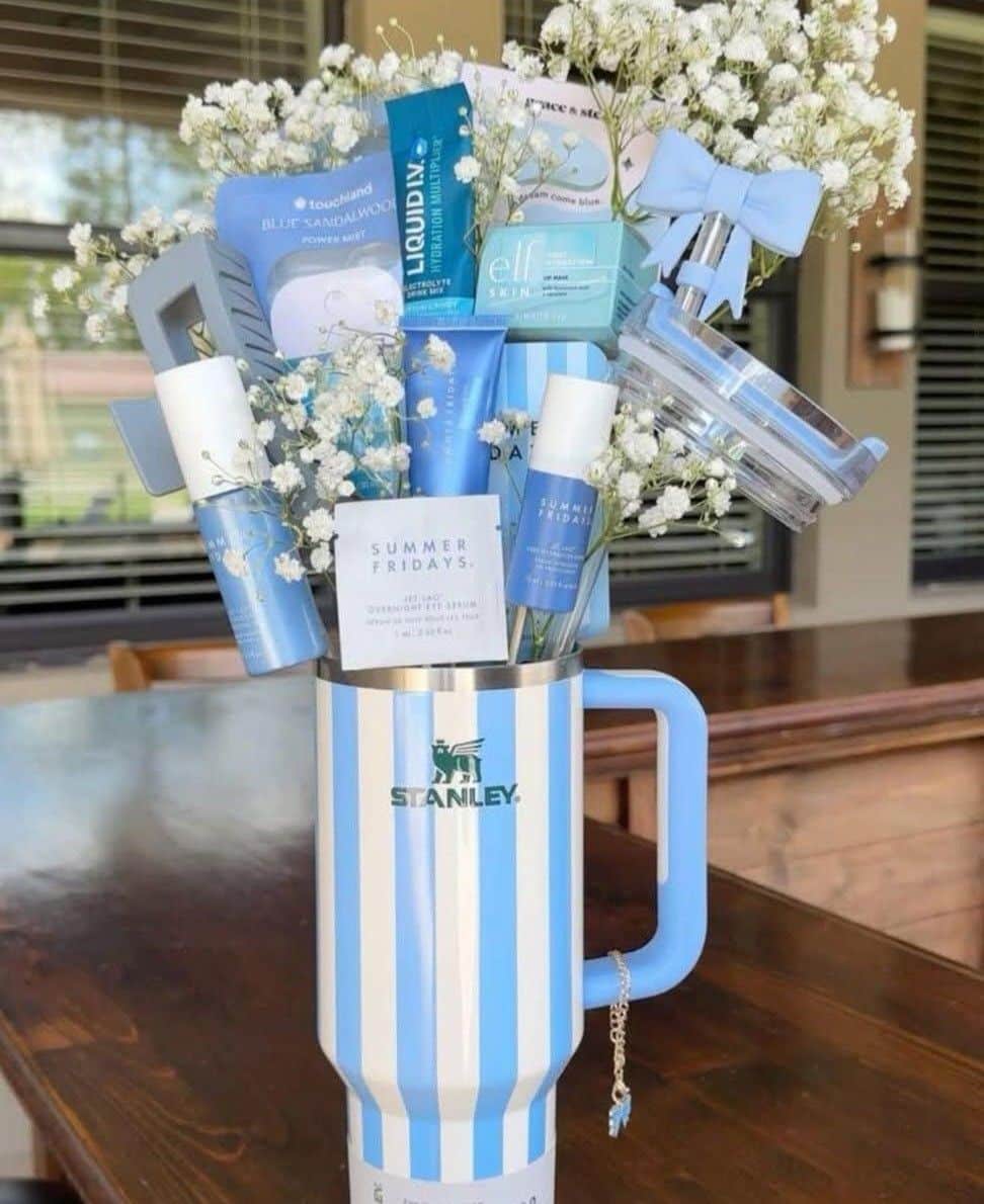 Blue and white striped Stanley tumbler on a wooden table filled with white baby's breath flowers, Summer Fridays skincare products, Touchland Blue Sandalwood hand mist, Liquid IV hydration, e.l.f. lip mask, and a charm bracelet