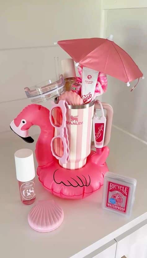 Pink striped Stanley tumbler sitting inside an inflatable flamingo pool float with a mini umbrella on top, Sol de Janeiro body mist, Aquaphor lip product, a makeup brush, and waterproof playing cards