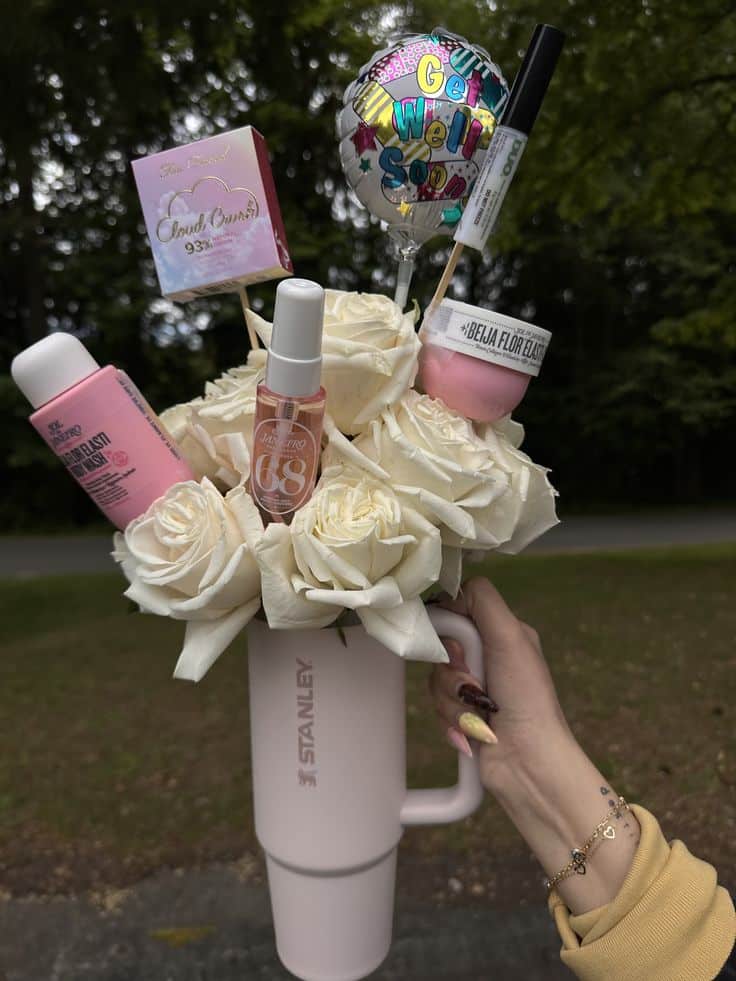 Pink Stanley tumbler held outdoors and filled with white roses, a Get Well Soon mini balloon, Too Faced Cloud Crush foundation, Sol de Janeiro body wash, Beija Flor body cream, and a nail serum