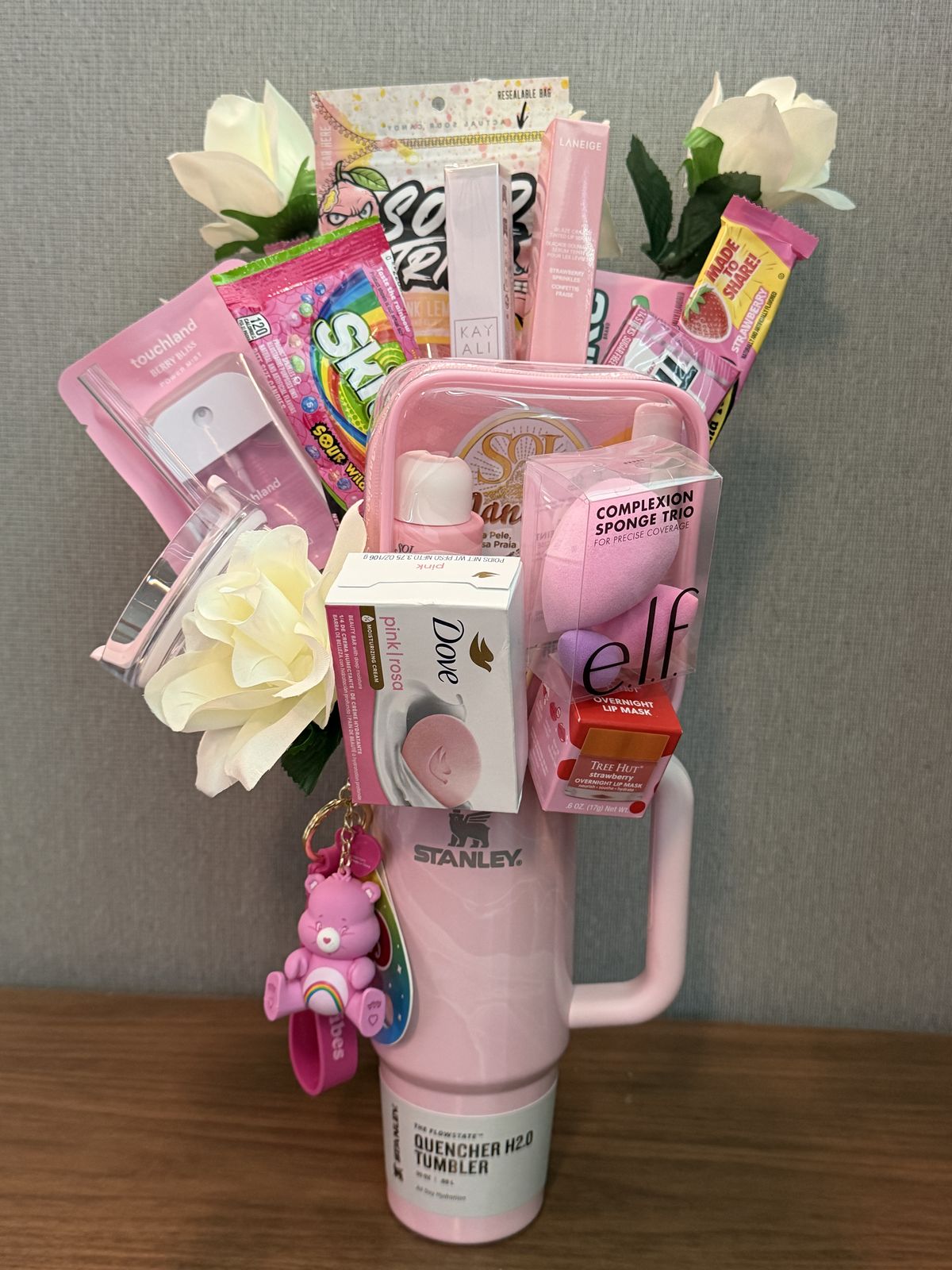 Pink Stanley tumbler decorated with Care Bear keychain and packed with e.l.f. beauty products, Laneige lip mask, Dove soap, Skittles candy, and artificial white roses