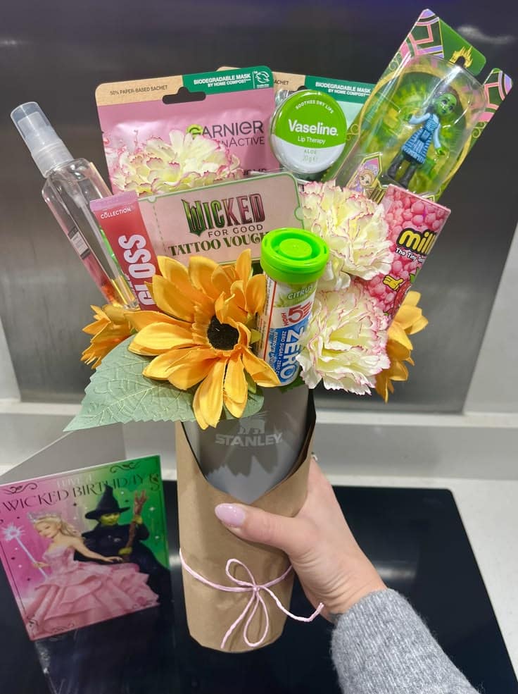 Black Stanley tumbler wrapped in brown paper and filled with sunflowers, cream carnations, a Wicked For Good tattoo voucher, Vaseline lip therapy, Garnier sheet mask, and candy