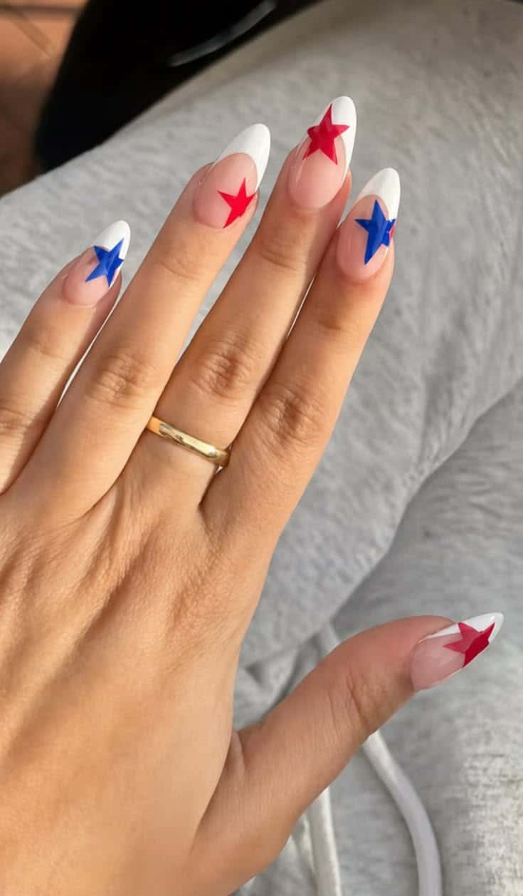 Long almond nails with a natural base and white French tips, each nail featuring either a red or blue star at the tip, photographed resting on a grey fabric background.