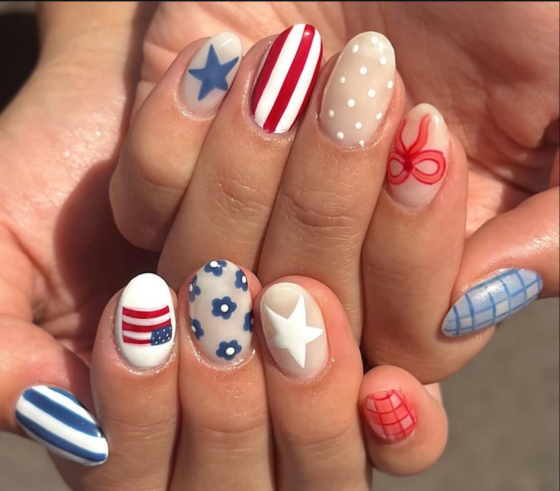Short oval nails featuring a maximalist patriotic nail art set with designs including a mini American flag, blue daisy florals on a nude base, red stripes, a large white star, a red bow outline, and blue and red gingham on each finger.
